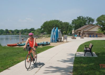A person wearing a pink helmet and orange top rides a bicycle on a lakeside path near a beach area with kayaks, a building, trees, and benches on a sunny day.