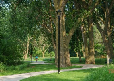 A winding paved trail passes through a green, tree-filled park on a sunny day. A person rides a bike in the distance, and benches and a lamppost line the pathway.