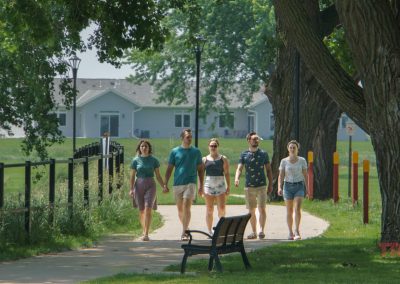Five adults walk together on a sunny, tree-lined path near houses. A black fence runs along the path, and a bench is in the foreground.
