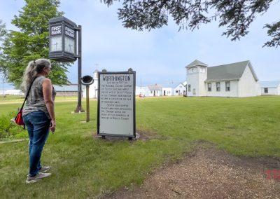 A woman with gray hair stands outdoors reading a historical sign for Worthington, MN, near an old-fashioned bank clock and a white building with a steeple on a grassy lawn.