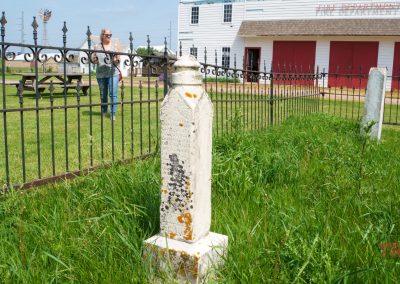 An old, weathered white gravestone stands in tall grass within a fenced cemetery. A woman in sunglasses walks nearby. In the background, there’s a white building with red doors and “FIRE DEPARTMENT” written on it.