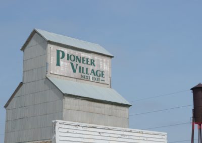 A tall, pale building with a sign reading Pioneer Village Next Exit stands against a blue sky. A rusted water tower is visible in the background.