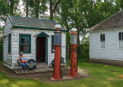 A woman sits on the porch of a small, weathered white building with green trim, next to two vintage red gas pumps; trees and another old white building are in the background.