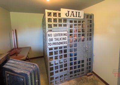 A small jail cell with barred walls and a sign reading NO LOITERING OR TALKING TO PRISONERS. The white JAIL sign is above the cell. The room contains folded tables and chairs.
