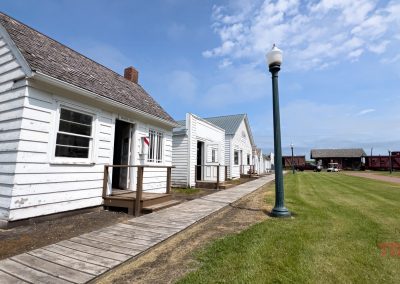 A row of small white wooden buildings with gabled roofs lines a wooden walkway. A vintage streetlamp stands on green grass under a blue sky with scattered clouds. In the distance, there are red train cars.
