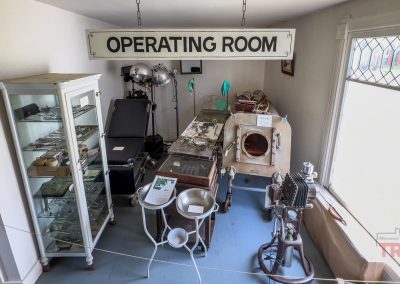 A small room displays vintage medical equipment, including an iron lung, glass cabinet with tools, examination tables, and surgical lamps, under a large sign reading “OPERATING ROOM.” Sunlight enters through a glass window on the right.