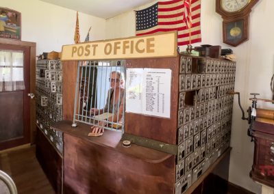 Old-fashioned post office with a person behind a barred counter, surrounded by vintage mailboxes. American flags, a wall clock, and antique items decorate the space, giving it a nostalgic atmosphere.