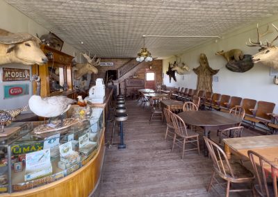 A vintage room with animal heads mounted on the walls, wooden chairs and tables, and a glass display case filled with antique items and bird taxidermy. The room has a rustic, old-fashioned atmosphere.