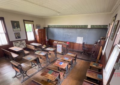 A vintage classroom with wooden desks, chalkboards, and educational posters. Sunlight enters through tall windows, and portraits hang on the walls. The atmosphere feels historical and preserved.