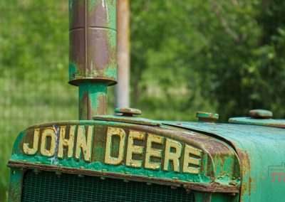 Close-up of the rusty front of an old green John Deere tractor, with faded yellow lettering and chipped paint, set outdoors with blurred green foliage in the background.