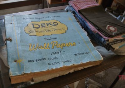 A large, worn book labeled Deko Products Decorators Wall Paper Co. De Luxe Wallpapers 1961 sits open on a glass counter next to folded fabrics and papers in a vintage store.
