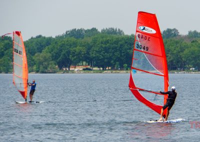 Two people windsurfing on a lake, each on a board with a brightly colored sail, with trees and buildings visible on the far shore under a clear sky.