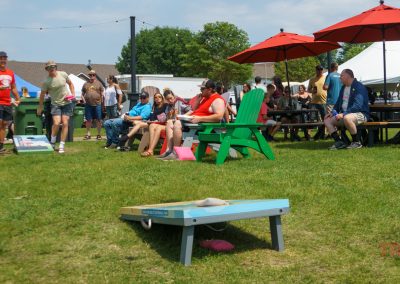 People at an outdoor event play cornhole on the grass, with some seated on colorful chairs and benches under red umbrellas. Others stand and watch. Its a sunny day with tents and houses in the background.