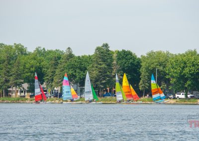 Several colorful sailboats with tall sails line up near the shore of a lake, with trees and houses in the background under a clear sky. The water is calm and the scene appears peaceful and lively.