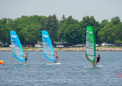 Three people windsurf on a calm lake with blue and green sails, near an orange buoy. Trees, parked cars, and cabins are visible along the shoreline in the background under a clear sky.