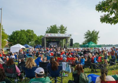 A large crowd sits in lawn chairs facing an outdoor stage at a lakeside park, enjoying a live music performance. Trees and vendor tents surround the area, with water visible in the background under a partly cloudy sky.