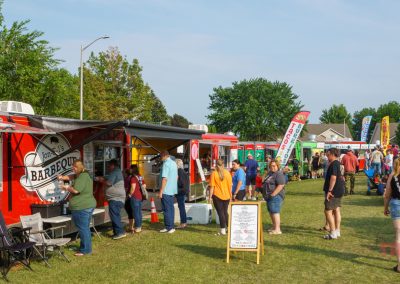People line up at colorful food trucks in a grassy park during a sunny outdoor festival. There are banners, trees, and groups of people walking, eating, and socializing near the trucks.