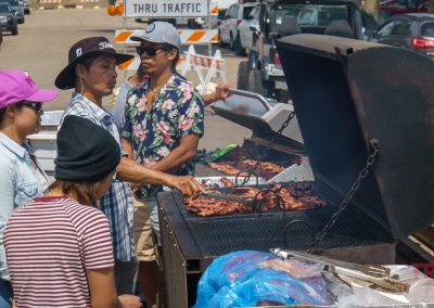 Several people gather around a large outdoor grill cooking meat. One man flips meat with tongs while others stand nearby, wearing hats and sunglasses. Cars and a Thru Traffic sign are visible in the background.
