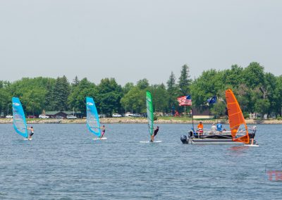 Windsurfers with colorful sails (blue, green, and orange) race near a boat with people and an American flag on a lake; trees and houses are visible along the shoreline in the background.