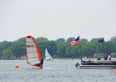 A person windsurfing near an orange buoy on a lake, with a boat displaying the American flag and several people onboard nearby. Houses, trees, and another sailboat are visible in the background.