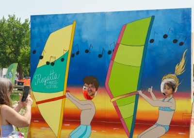 A woman takes a photo of two children posing with their faces in a painted cutout of windsurfers at the Regatta Music Festival, with a lake and grassy area in the background.