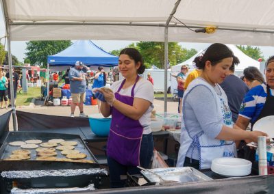 Three women prepare food at an outdoor festival booth, cooking tortillas or pupusas on a griddle under a white tent. People and colorful tents are visible in the background on a sunny day.