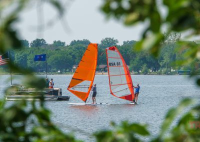 Two people windsurf on a lake with bright orange sails, framed by green leaves. An American flag and another boat are visible on the left, and the shoreline with trees is in the background.