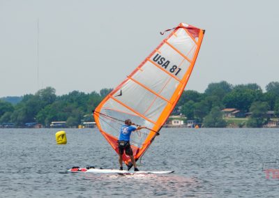 A person windsurfing on a lake, wearing a blue shirt and black shorts. The sail is orange and white with USA 81 printed on it. There is a yellow buoy on the water and houses with trees along the shoreline.