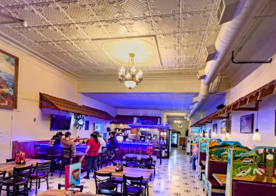 A vibrant restaurant interior with ornate ceiling, chandelier, wooden tables, colorful booths, and people ordering at the counter; walls decorated with paintings and warm lighting throughout.