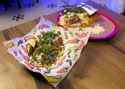 A colorful basket of tacos with chopped meat, onions, cilantro, and lime sits on a table, with a plate of rice, beans, tacos, and foil-wrapped food in the background.
