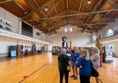 A group of four people stand talking inside a spacious gymnasium with wooden floors and a high vaulted ceiling. Large decorative skeleton figures are displayed, and exhibit panels line the walls. Natural light streams through arched windows.