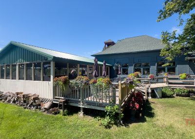 A rustic restaurant with green roofs, outdoor seating, potted flowers, and a woodpile, set on a grassy lawn under a clear blue sky.