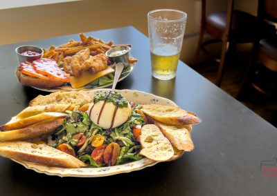 A table with a plate of salad topped with burrata, cherry tomatoes, and toasted bread slices, alongside a basket of fries with sauce and a partially filled glass of beer.