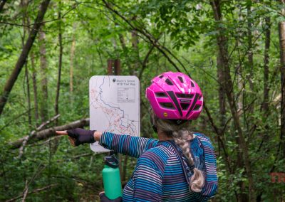 A person wearing a pink bike helmet and striped shirt points at a trail map posted on a sign in a lush green forest. A green water bottle hangs from their backpack.