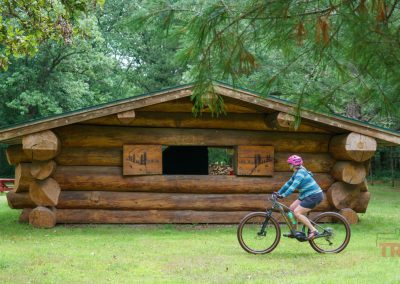 A person wearing a helmet rides a bicycle past a rustic log cabin in a grassy, wooded area. A red picnic table sits to the left, and trees surround the scene.