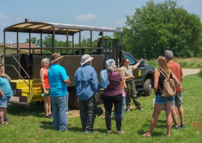 A group of people stands in a semicircle around a park ranger near an open-sided tour vehicle on a grassy field under a clear sky. The ranger appears to be explaining something to the group.
