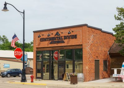 A small brick building with a sign reading Continental Divide Bar & Eatery sits on a street corner. A stop sign, American flag, and large decorative chair are nearby. Trees and other buildings are in the background.