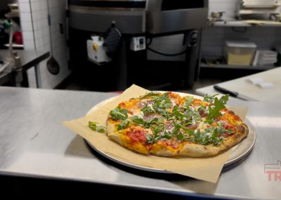 A freshly baked pizza topped with arugula, red onions, and tomatoes sits on parchment paper on a metal tray in a commercial kitchen.