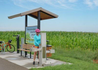 A person in a pink helmet and blue shirt uses a bike repair station at a rest stop beside a cornfield. An orange bicycle is parked nearby on a paved trail under a small shelter with a bench.