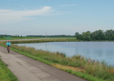A person in a blue top and helmet rides a bicycle on a paved path beside a calm, tree-lined lake under a partly cloudy sky; another cyclist is visible in the distance.