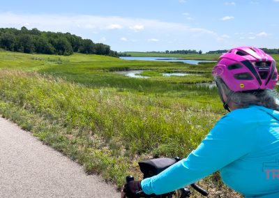 A person wearing a pink helmet and turquoise jacket rides a bike on a paved trail through green fields, with a small lake and trees in the background under a blue sky.