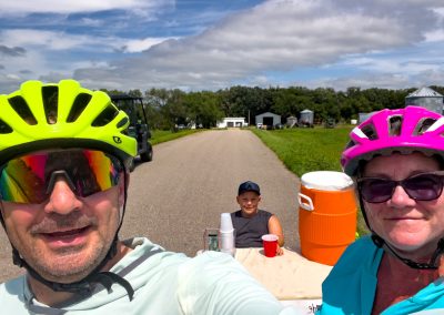 Two cyclists wearing bright helmets and sunglasses take a selfie on a rural road, with a boy sitting at a table with an orange drink cooler and cups behind them. Green fields and farm buildings are in the background.