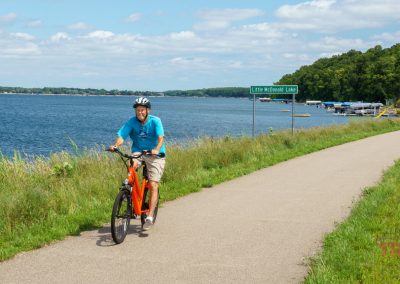 A person wearing a helmet rides a bicycle on a paved lakeside trail with grass on both sides. A sign reads Little McDonald Lake, and boats are visible on the water under a partly cloudy sky.