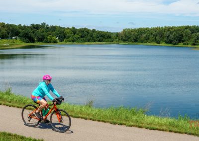 A person in a pink helmet and blue shirt rides a bike on a paved trail beside a large lake, surrounded by green trees and grass under a partly cloudy sky.