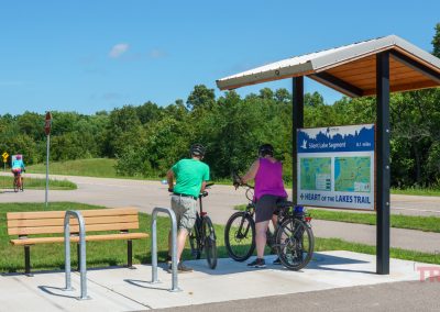Two cyclists stop at a shaded trail sign and map along a paved path. A bench and bike rack are nearby, with trees and another cyclist in the background under a blue sky.