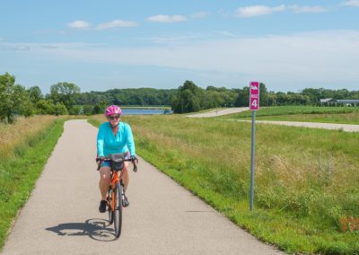 A person wearing a pink helmet and blue top rides a bicycle on a paved trail through a green, grassy landscape. A mile marker sign reads Mile 4. Trees and a blue sky with clouds are in the background.