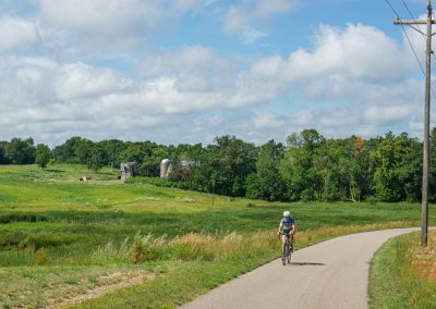 A person wearing a helmet and cycling gear rides a bicycle on a paved country road, surrounded by green fields, trees, and a partly cloudy sky. Power lines and a small barn are visible in the background.