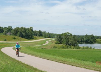 A person wearing a pink helmet and blue shirt rides a bicycle on a winding paved trail through green fields, with trees and a lake in the background under a partly cloudy sky.