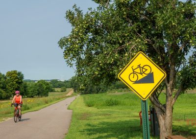 A cyclist rides on a paved trail surrounded by greenery. A yellow bike warning sign is posted on a pole next to a tree, indicating a downhill slope ahead. The sky is clear and the scene is bright and sunny.