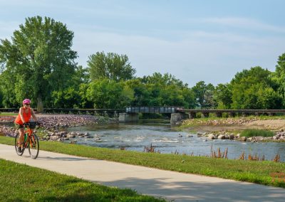 A person wearing an orange shirt and pink helmet rides a bicycle on a paved path next to a river, with a metal bridge and lush green trees in the background on a clear day.